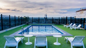 Ein Pool auf dem Dach mit Blick auf die Stadtlandschaft, umgeben von weißen Liegestühlen und Tischen.