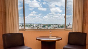 Una habitación con dos sillas y una mesita, una ventana con vistas a la ciudad y una botella sobre la mesa.