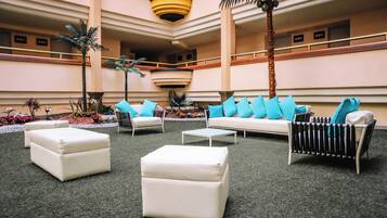 A spacious indoor area with a central seating arrangement, including a sofa and ottomans, surrounded by potted plants and a palm tree.