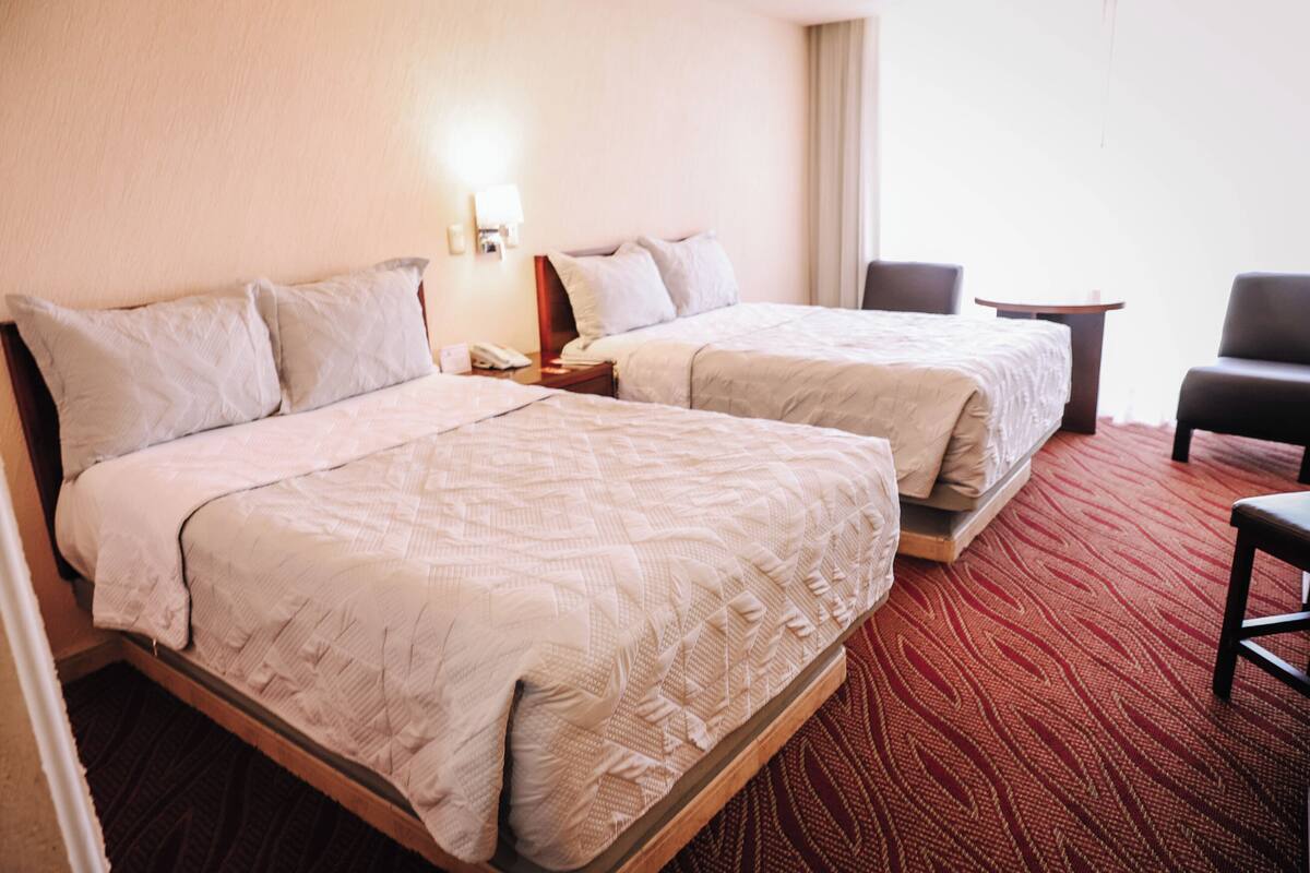 A hotel room with two beds, a small table, a chair, and a nightstand with a lamp.