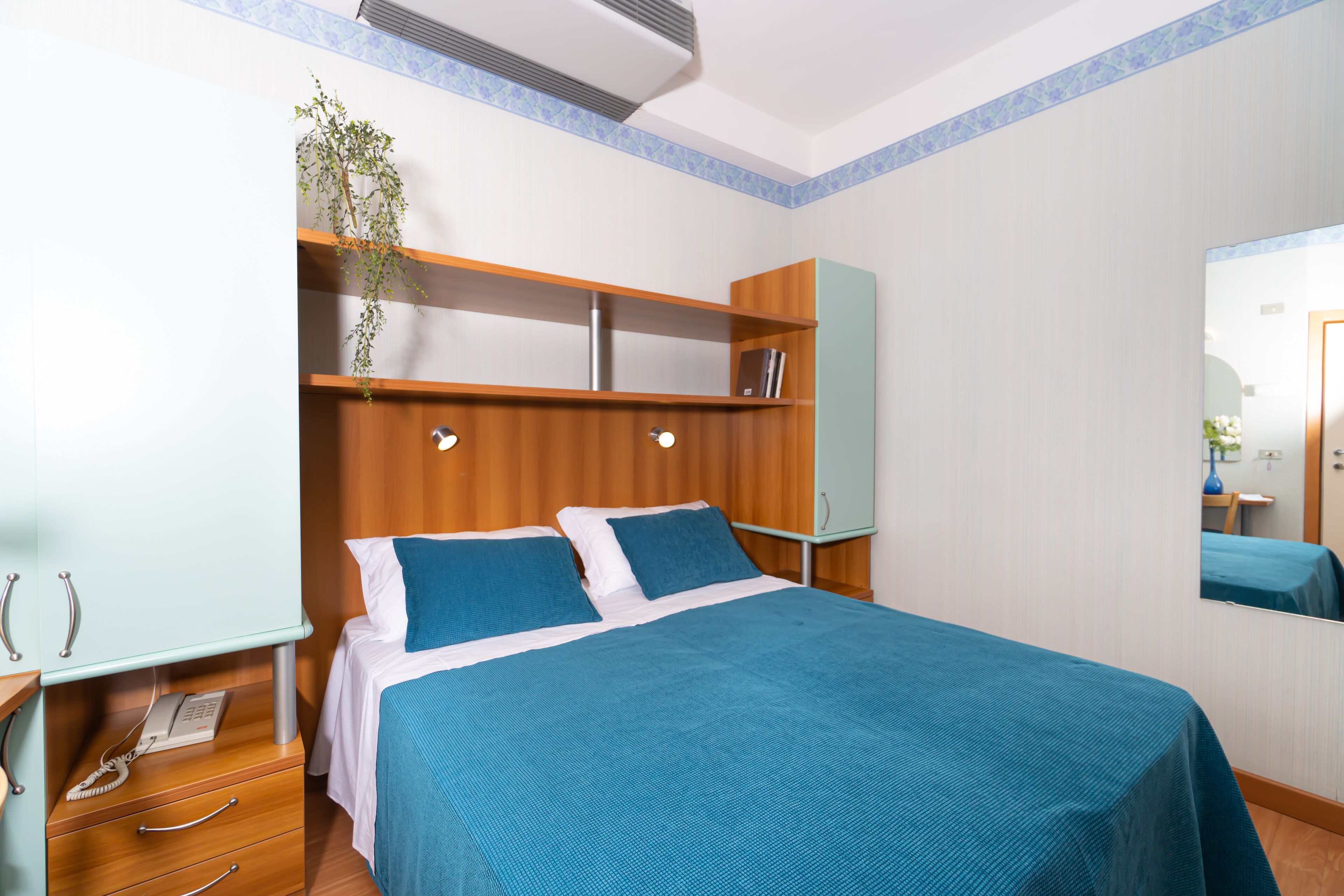 Habitación de hotel con cama, mesita de noche, espejo y estante con una planta.