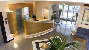 A reception desk with a modern design, a large mirror, and a potted plant. There is a seating area with a blue sofa and a small table. The floor has a geometric pattern. - Hotel Sampaoli (Bellaria-Igea Marina)