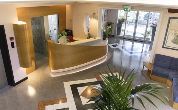 A reception desk with a modern design, a large mirror, and a potted plant. There is a seating area with a blue sofa and a small table. The floor has a geometric pattern. - Hotel Sampaoli (Bellaria-Igea Marina)