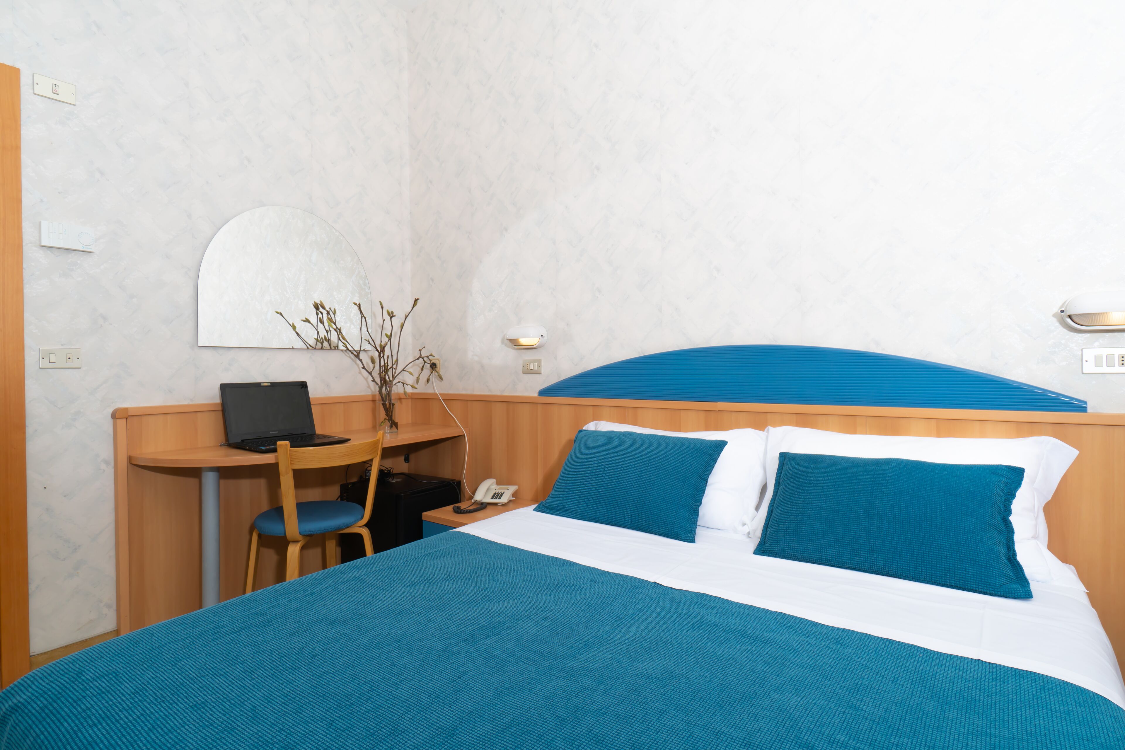 Quarto de hotel com cama, escrivaninha, cadeira, laptop e telefone.
