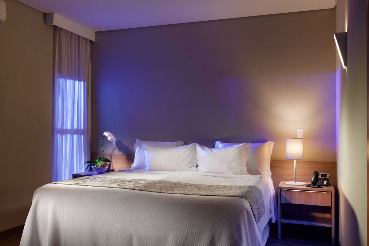 Habitación de hotel con una cama bien hecha, una mesita de noche con lámpara y una planta en maceta.