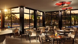 A spacious outdoor dining area with multiple tables and chairs, large windows offering views of palm trees and a pool, and ceiling fans with flower-shaped lights. - Malai Manso Yacht Convention & Spa - All inclusive (Chapada dos Guimarães)