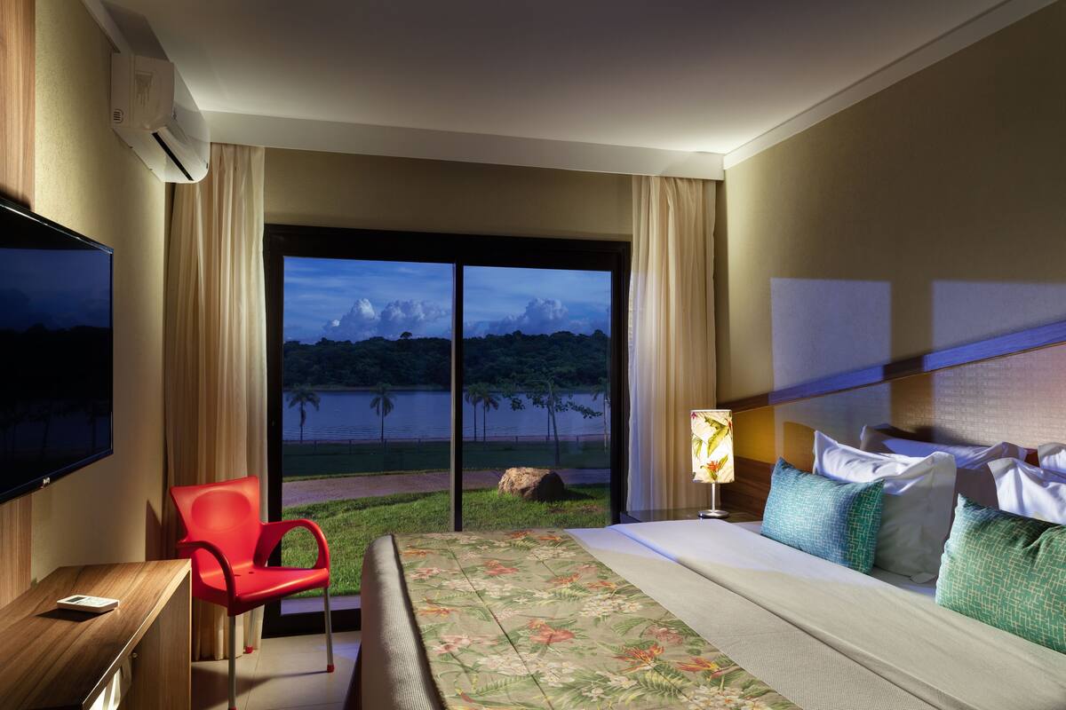 Habitación de hotel con cama, escritorio, televisor y ventana con vista al agua y árboles.