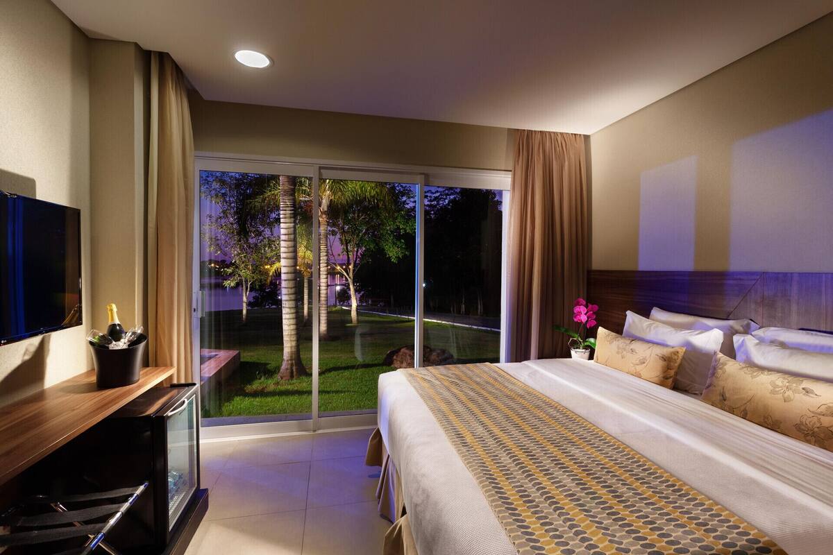 Habitación de hotel con una cama grande, televisión, minibar y puerta corrediza de vidrio que da a un jardín.