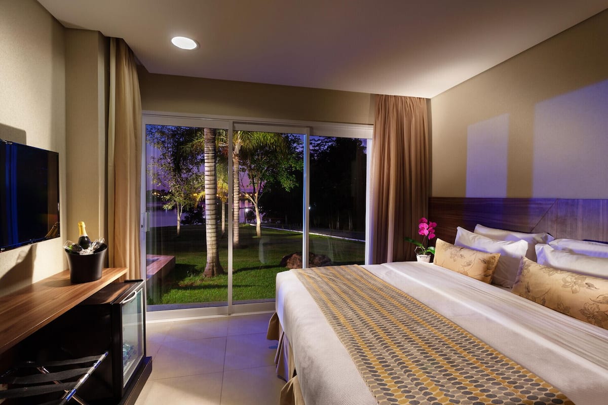 A hotel room with a large bed, a TV, a minibar, and a sliding glass door leading to a garden.