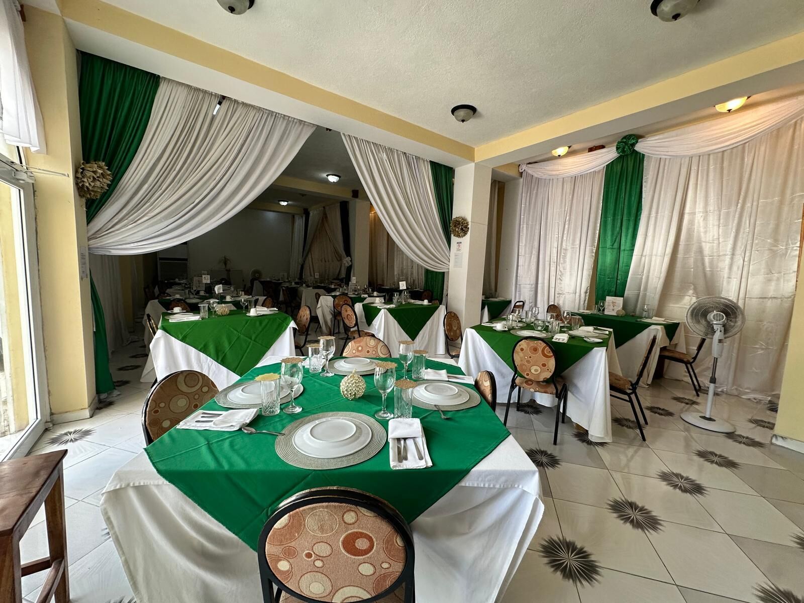 A dining area with tables set for multiple guests, featuring green tablecloths and white drapes.