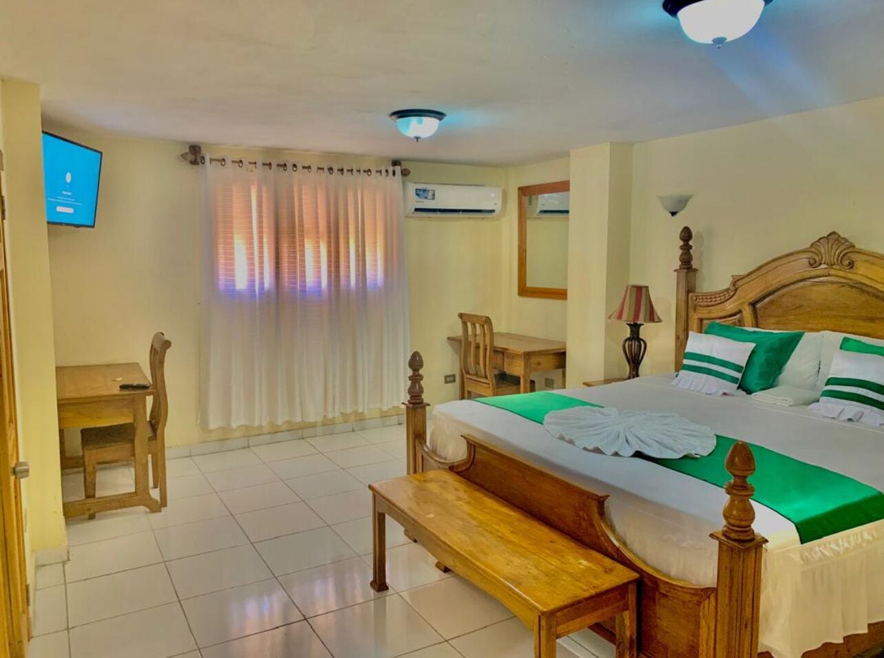 A hotel room with a wooden bed, a desk, a chair, a TV, and an air conditioning unit.