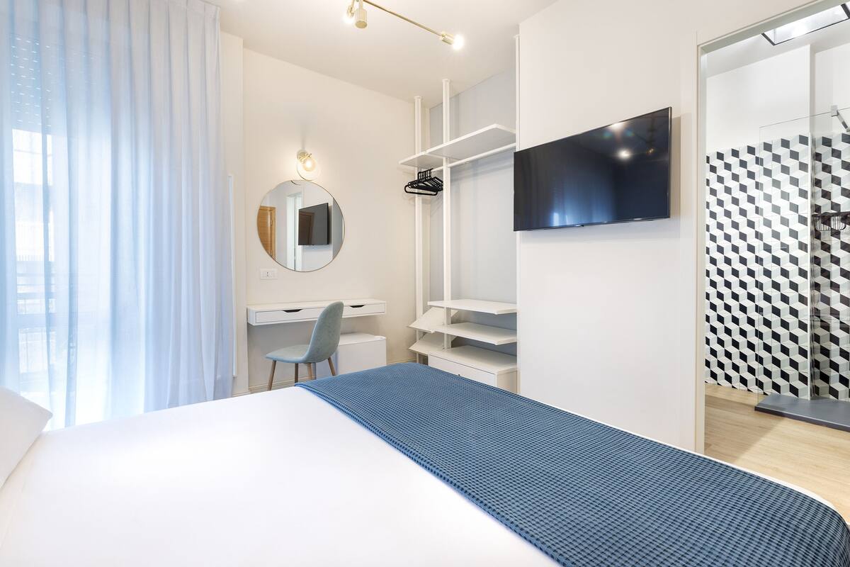 Habitación de hotel moderna con cama, escritorio, silla, televisor y zona de ducha.