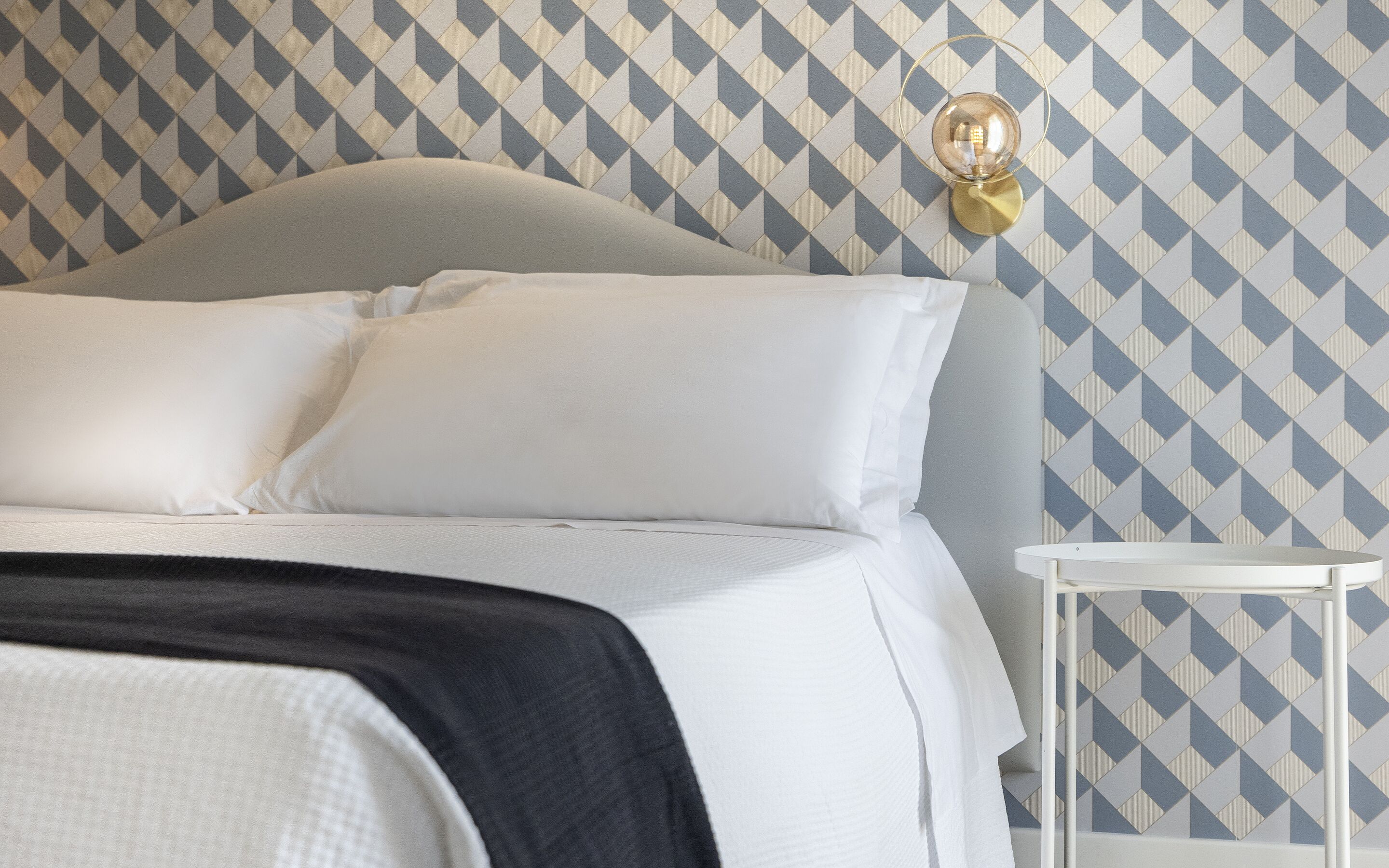 Un lit bien fait, avec du linge de lit blanc et un plaid noir, une petite table de chevet avec une lampe blanche, et un papier peint à motifs avec un dessin géométrique.