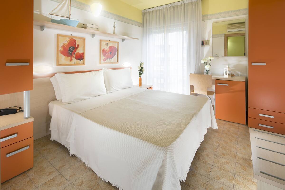 Un dormitorio con una cama grande, una mesita de noche, un aparador, un espejo y una ventana con cortinas.