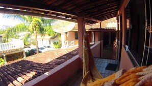 Una hamaca en un balcón con vista a un tejado de baldosas, palmeras y un edificio con tejado a dos aguas.