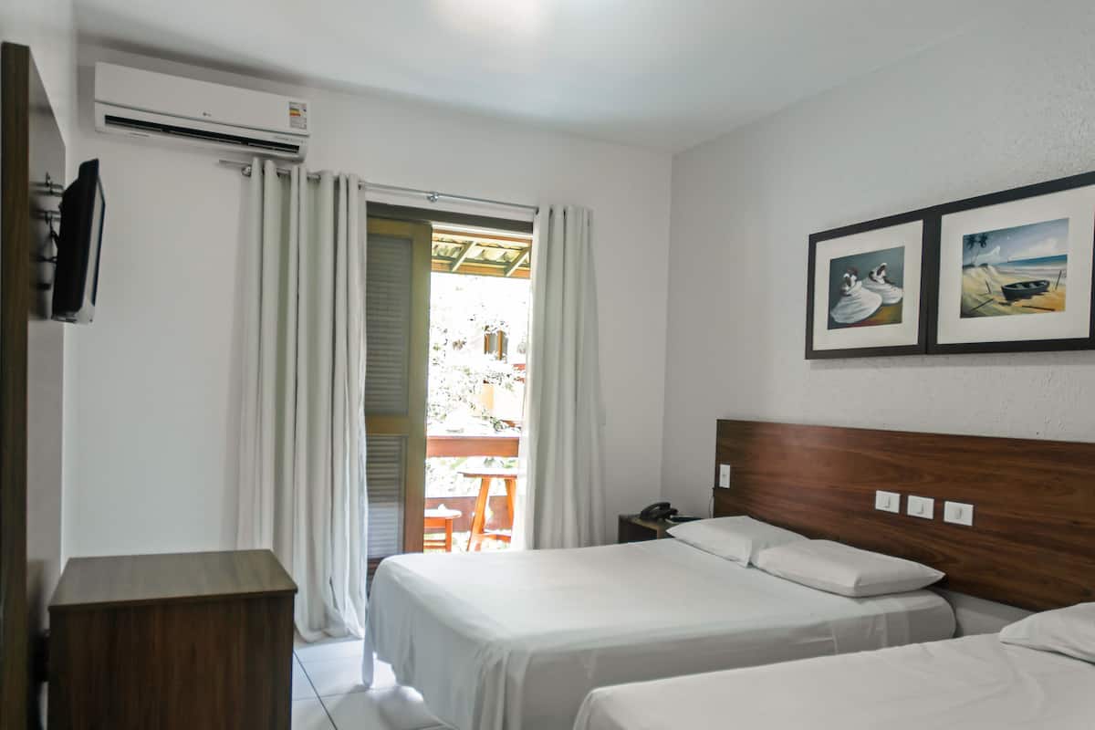 Une chambre d’hôtel avec deux lits, une tête de lit en bois, un bureau, un climatiseur, une fenêtre avec des rideaux et deux tableaux encadrés au mur.