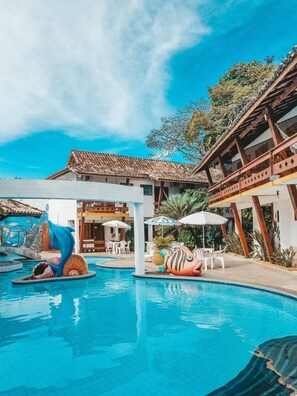 A swimming pool with dolphin and fish sculptures, surrounded by outdoor seating and a building with a tiled roof. - Casa Blanca Park (Porto Seguro)