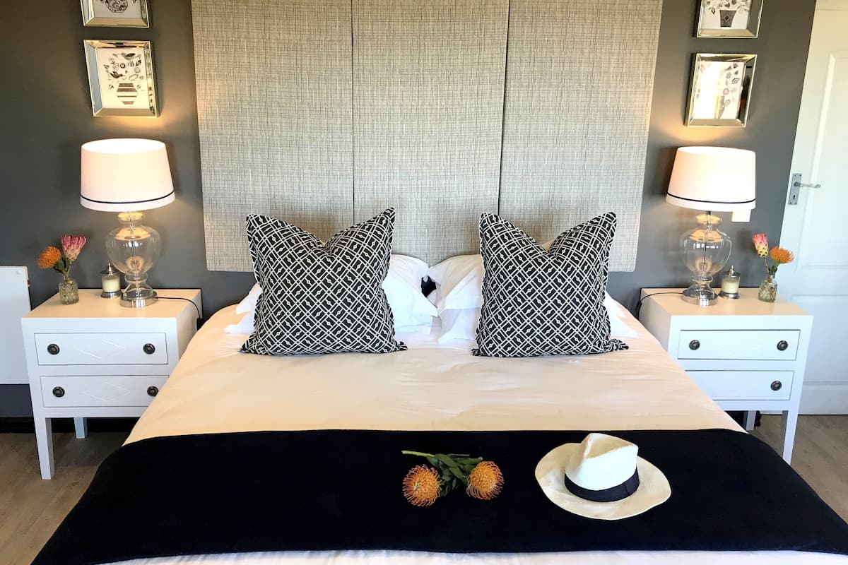 Una cama bien hecha con dos almohadas de diseño blanco y negro, una manta negra y un sombrero blanco sobre la cama. Hay dos mesitas de noche con lámparas y pequeños jarrones con flores. La habitación tiene una pared con textura e imágenes enmarcadas sobre la cama.