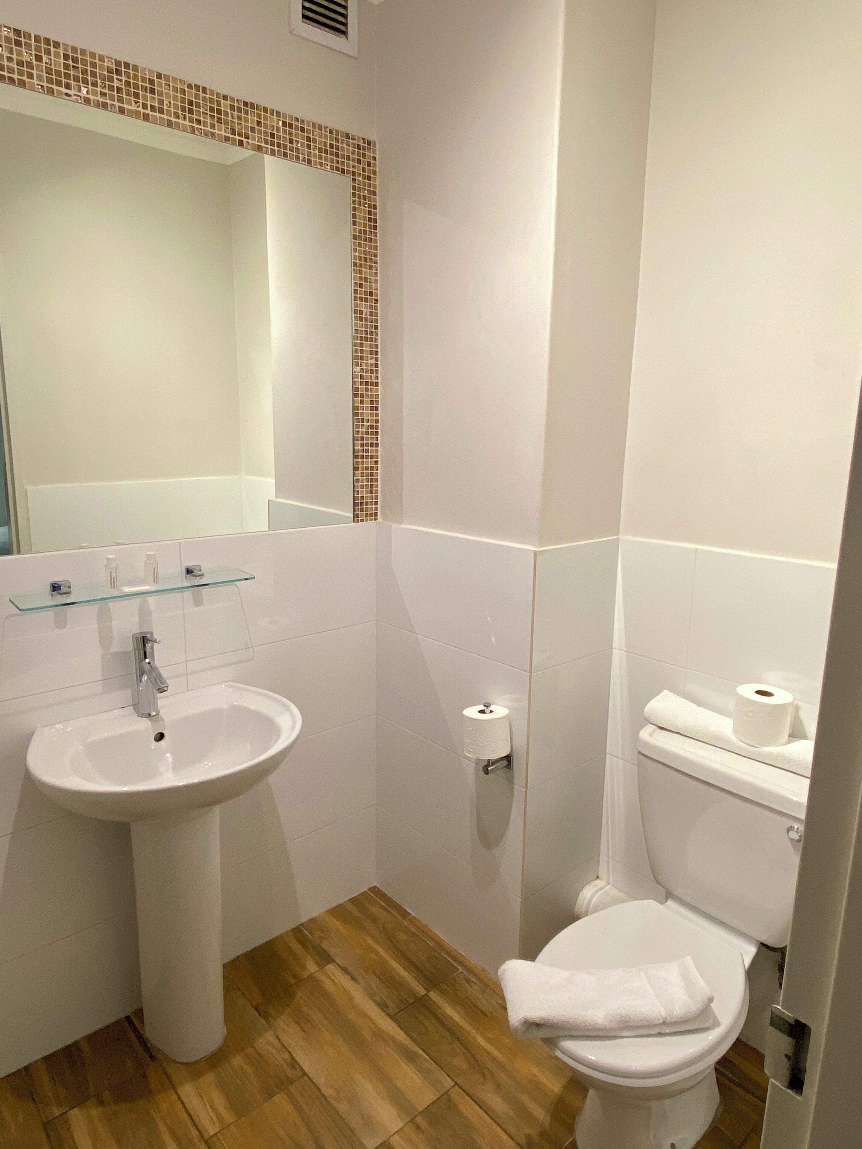 A bathroom with a white sink, a mirror with a mosaic border, a toilet, and a wooden floor.