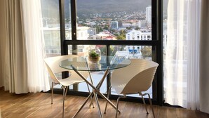 A room with a glass table and two chairs, a small plant on the table, and a view of a cityscape with a mountain in the background.