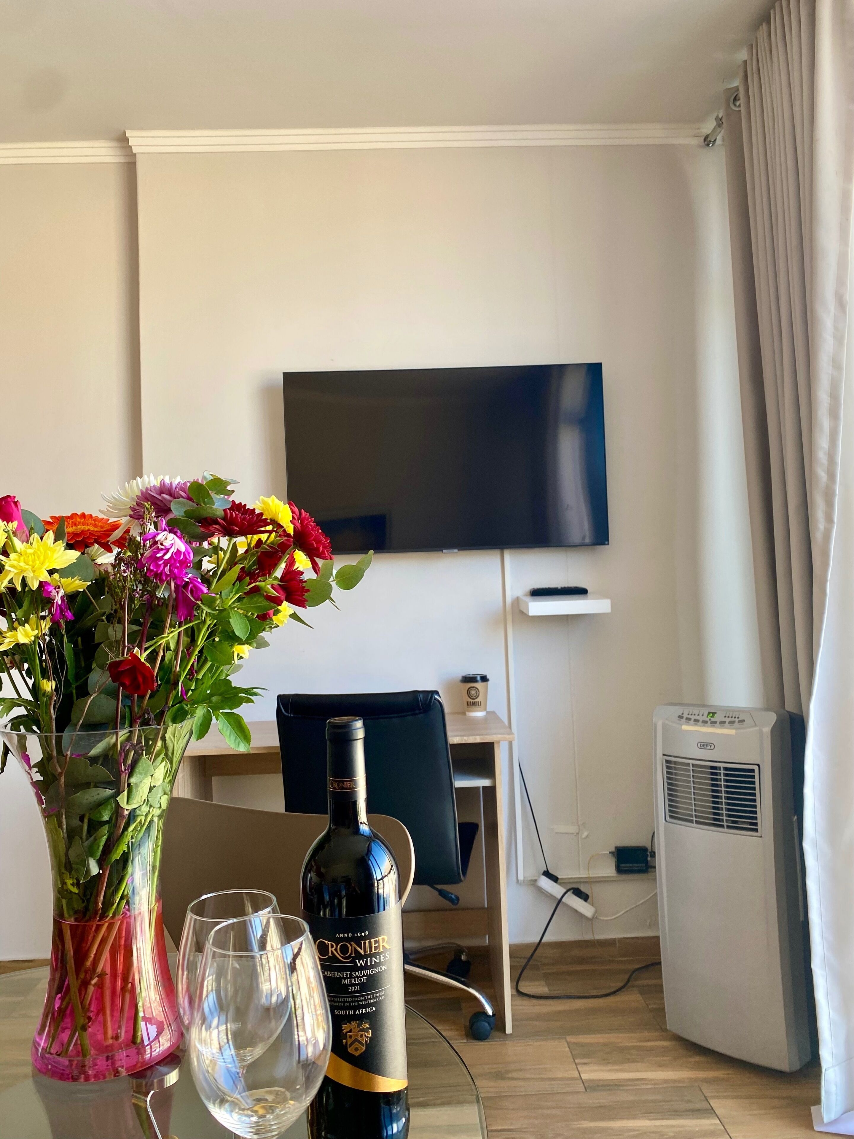 Ett hotellrum med en TV som är monterad på väggen, ett skrivbord med en stol, en minibar och ett fönster med gardiner.