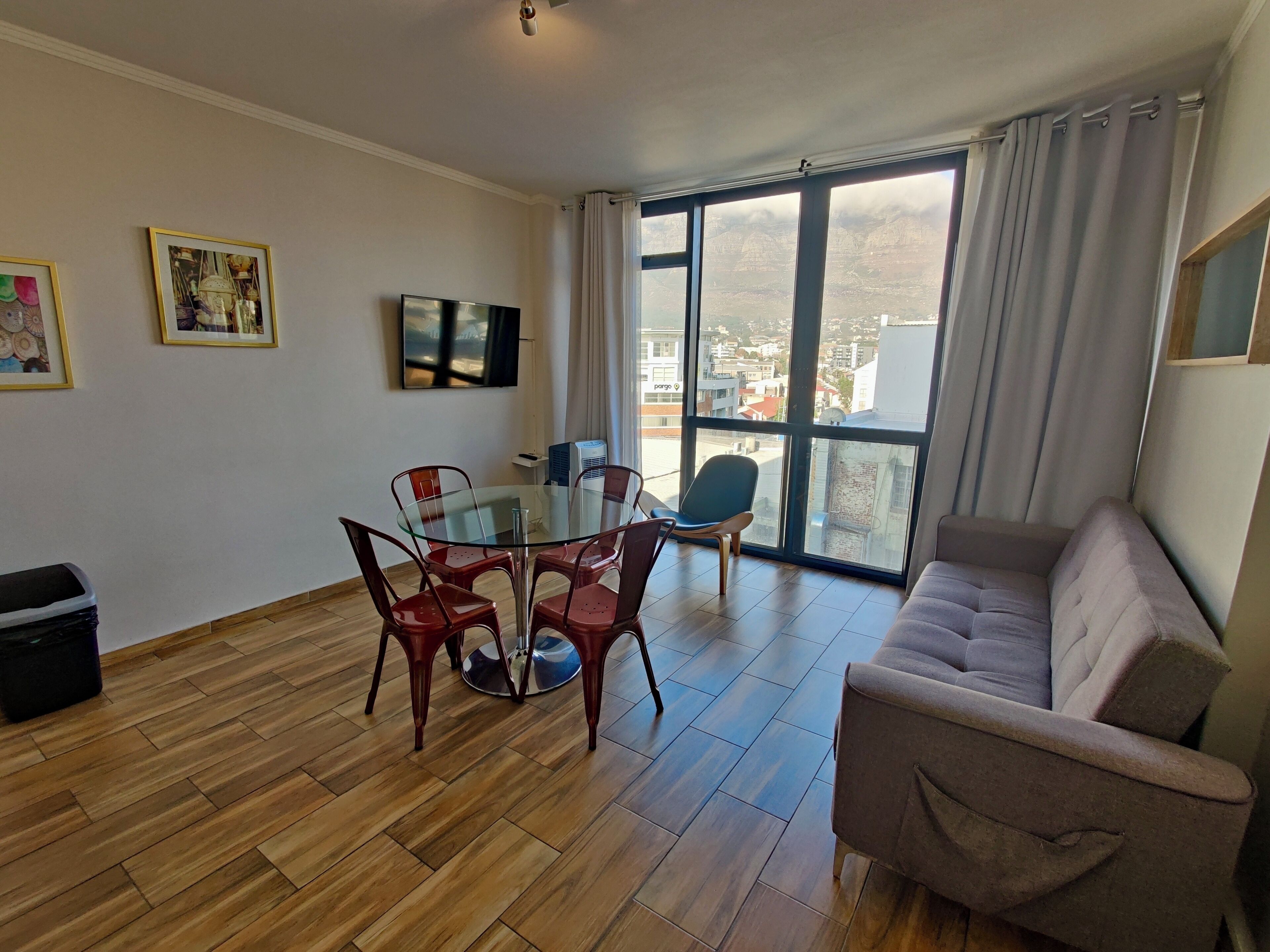 A modern living room with a glass dining table, red chairs, a grey sofa, and a large window offering a city view.