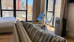 A hotel room with a large window, a bed, a sofa with patterned cushions, a small table with a plant, and a standing air conditioner.