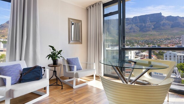 A modern living room with a glass table, two sofas, a small side table with a plant, and a view of a mountainous landscape.