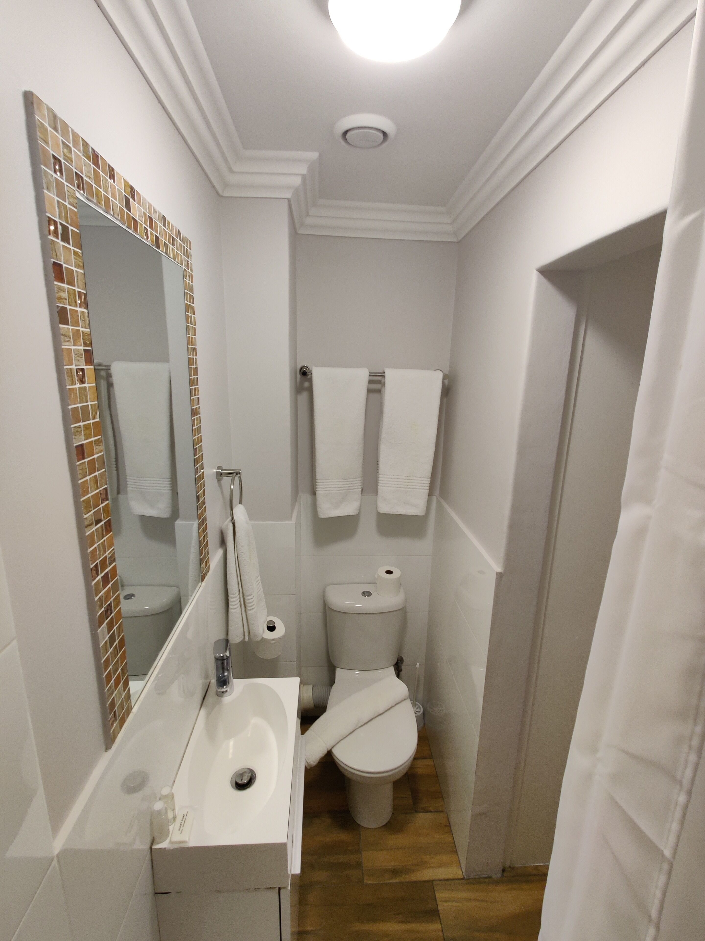 Un baño pequeño con lavamanos blanco, inodoro y espejo con zócalo de mosaicos.