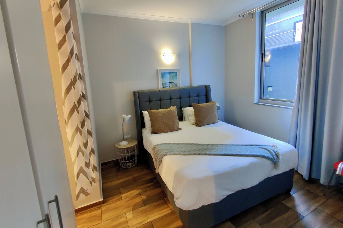 Une chambre à coucher comprenant un lit, une table de chevet, une lampe, une fenêtre avec des rideaux et un luminaire fixé au mur.