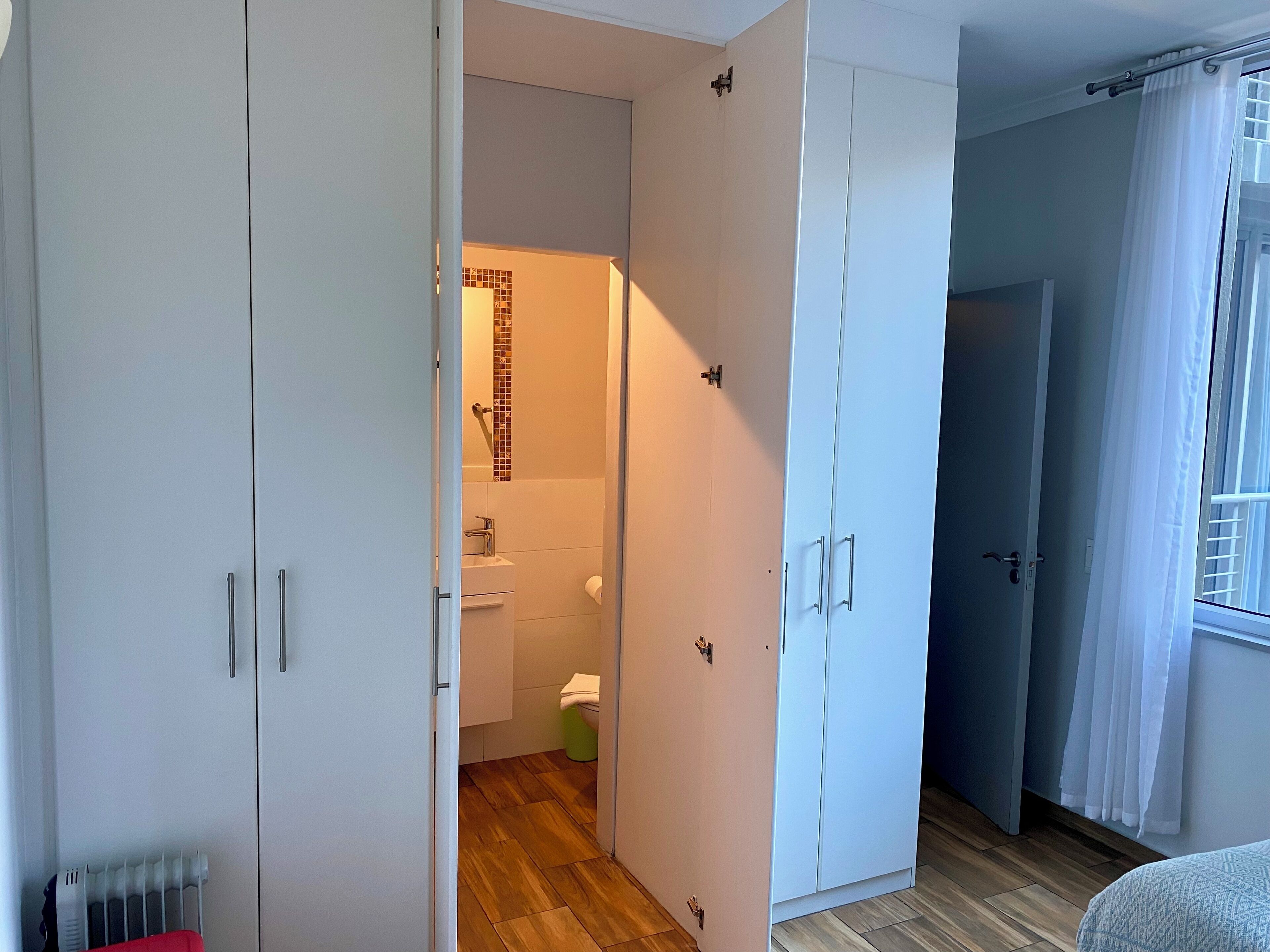 Un dormitorio moderno con un armario grande, un baño pequeño con ducha y una ventana con cortinas.