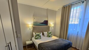 A hotel room with a large bed, two bedside tables, a nightstand, a painting on the wall, and a window with curtains. - Urban Oasis at Perspectives (Cape Town)