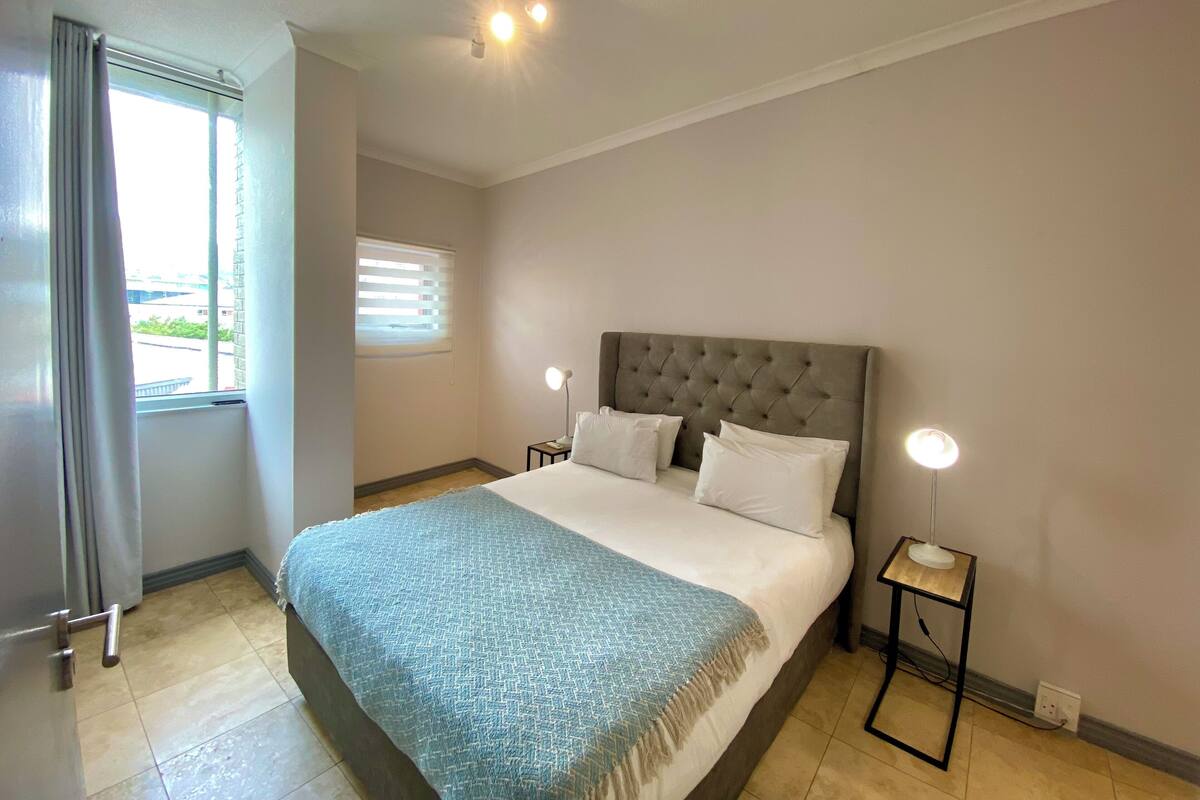 Une chambre à coucher comprenant un lit, deux tables de chevet, une fenêtre avec des stores et un luminaire au plafond.