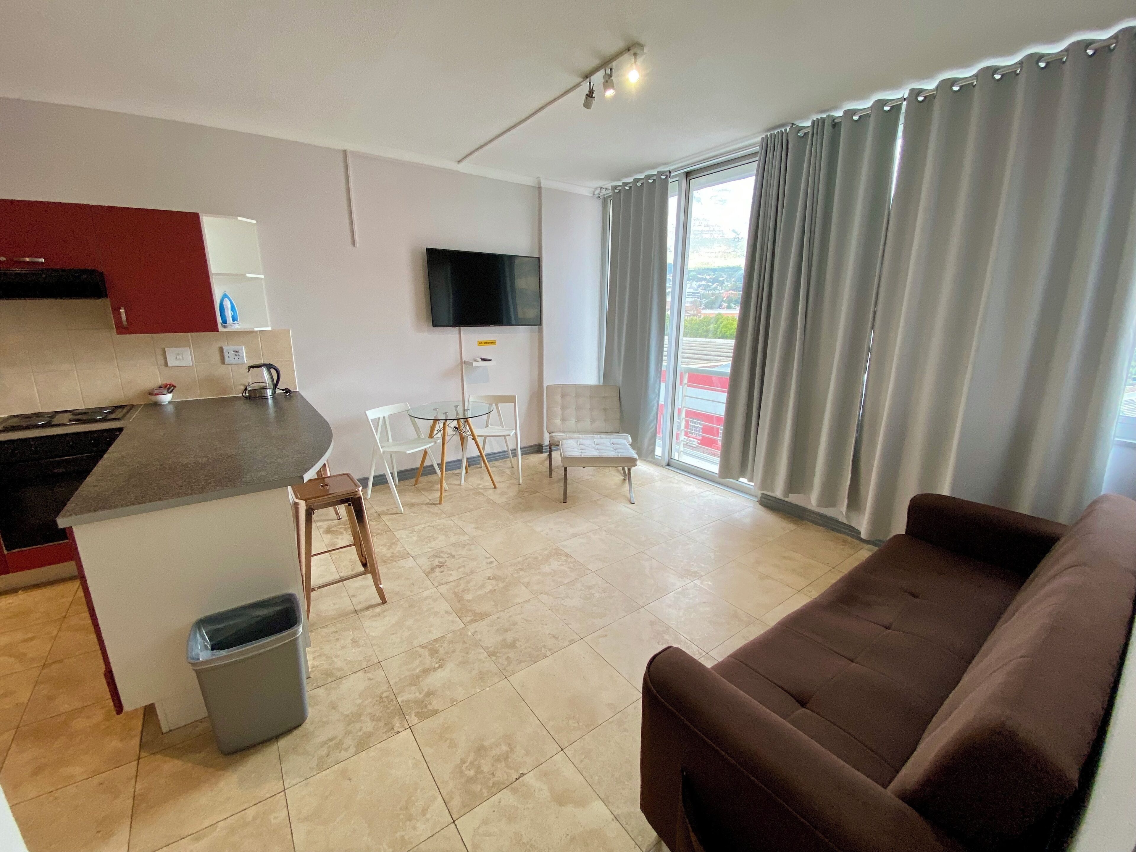 A modern living room with a brown sofa, a kitchen area with a stove, a TV, and a dining table with chairs.