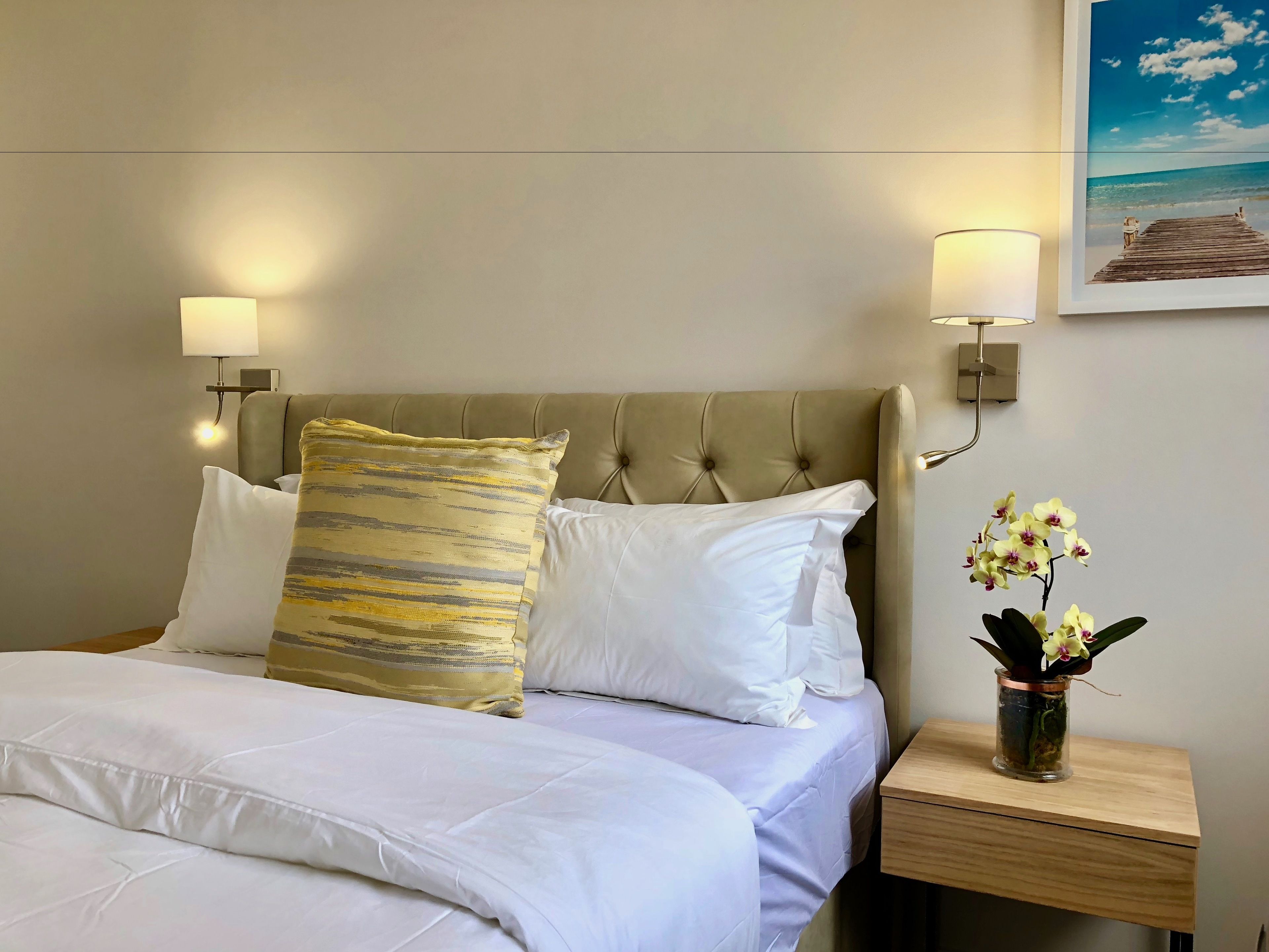 A neatly made bed with white linens and a patterned pillow, a wooden nightstand with a vase of orchids, and a wall-mounted lamp.
