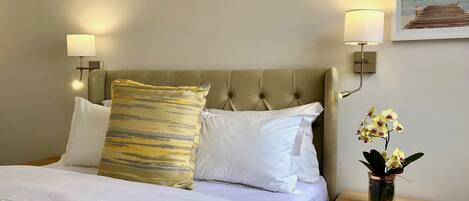 A neatly made bed with white linens and a patterned pillow, a wooden nightstand with a vase of orchids, and a wall-mounted lamp.