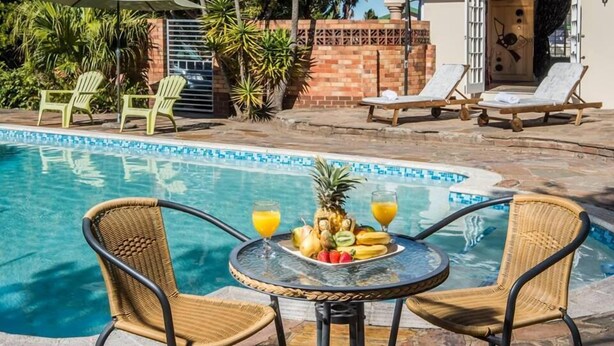 A poolside area with lounge chairs, a table with fruit and drinks, and a building entrance. - Bhotani Bed and Breakfast (Gqeberha (Port Elizabeth))