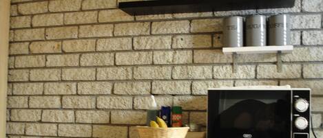 Una cocina con pared de ladrillo, un microondas, un refrigerador, un estante con latas y una cesta con limones.