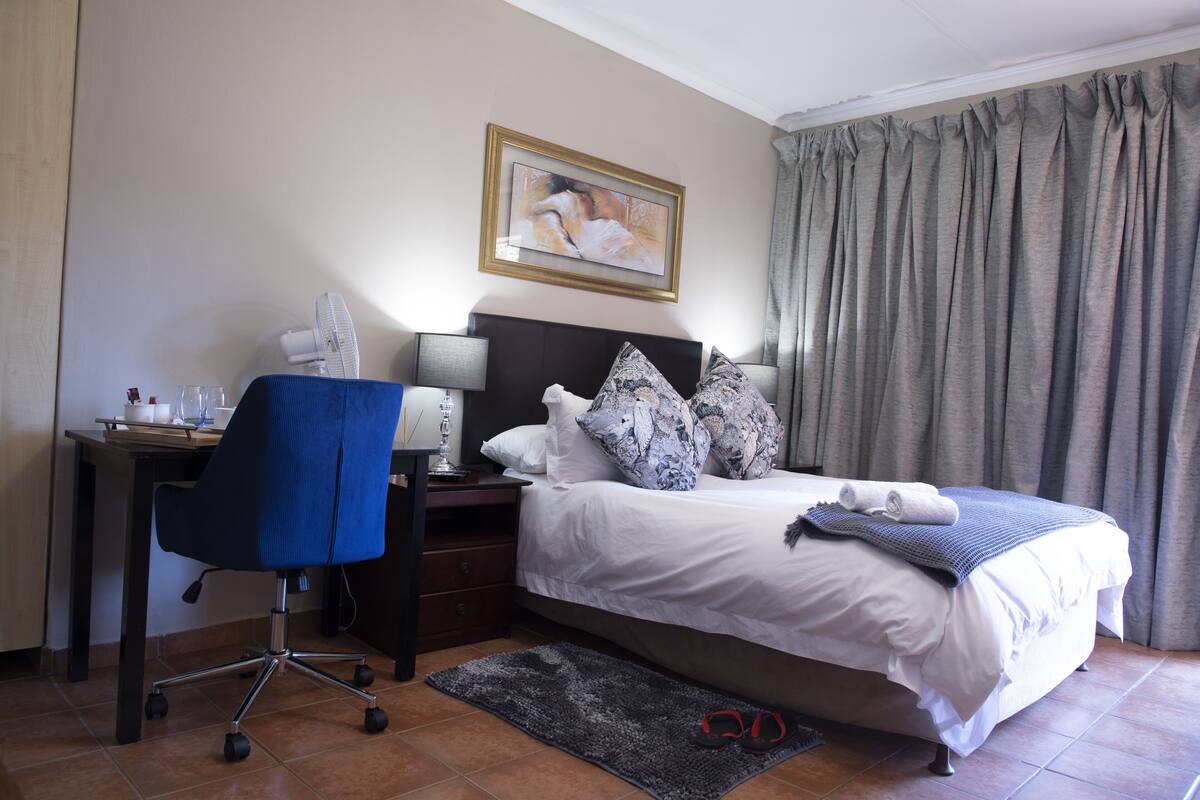 A hotel room with a bed, a desk with a chair, a nightstand, a framed picture on the wall, and a window with curtains.