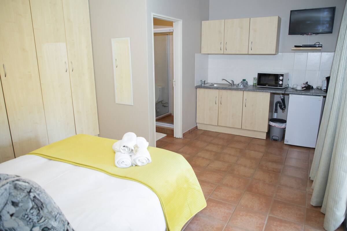 A hotel room with a bed, a yellow bedspread, a small kitchen area with a microwave, and a TV on the wall.