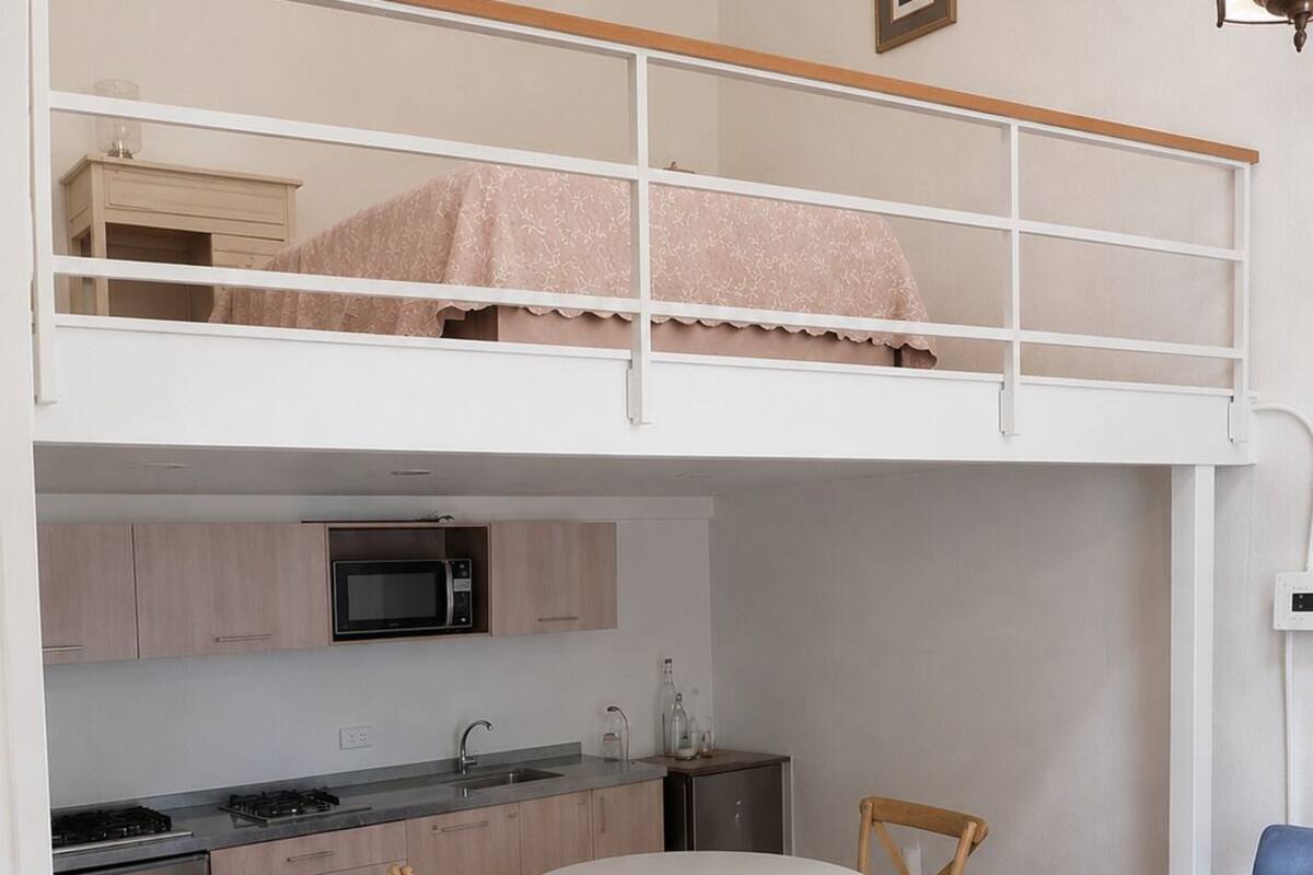 A loft-style bedroom with a bunk bed, a kitchenette with modern appliances, a dining area with a round table and chairs, and a blue sofa.
