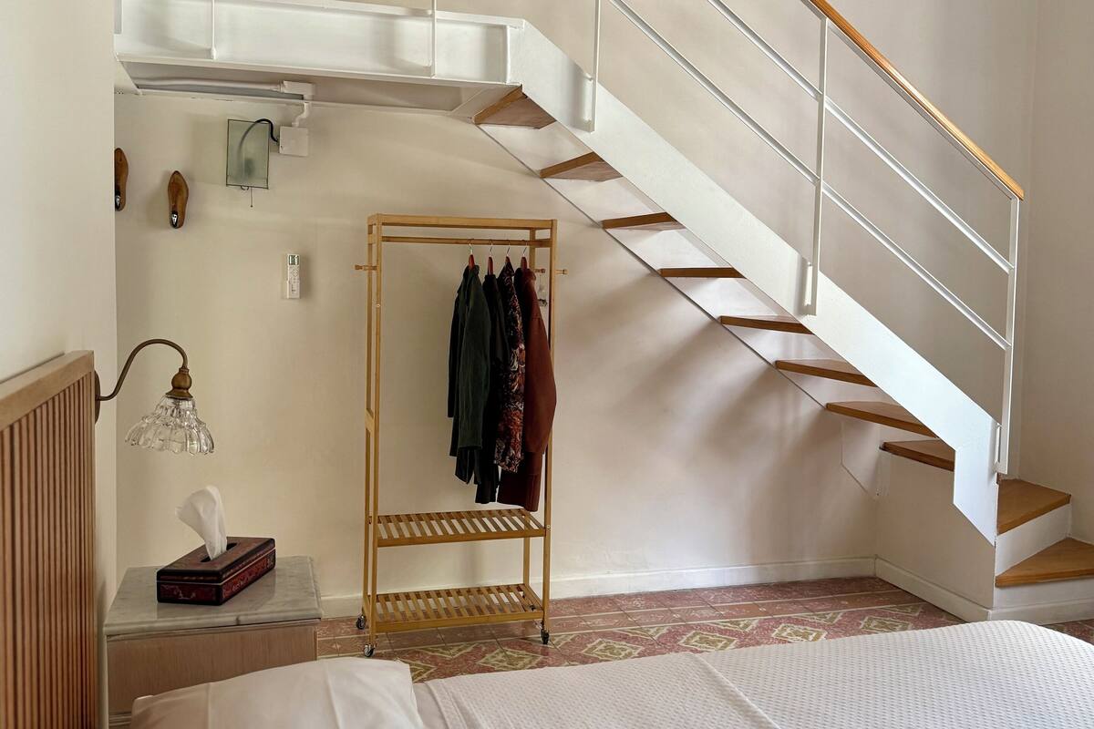 A bedroom with a bed, a wooden nightstand, a bedside table with a lamp, a hanging clothes rack, and a staircase with a wooden handrail.