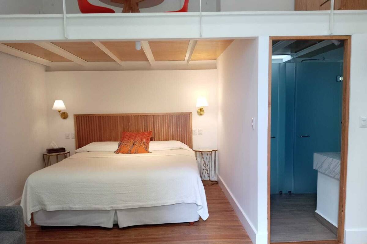 A bedroom with a large bed, wooden headboard, bedside lamps, and a view of the bathroom through an open door.