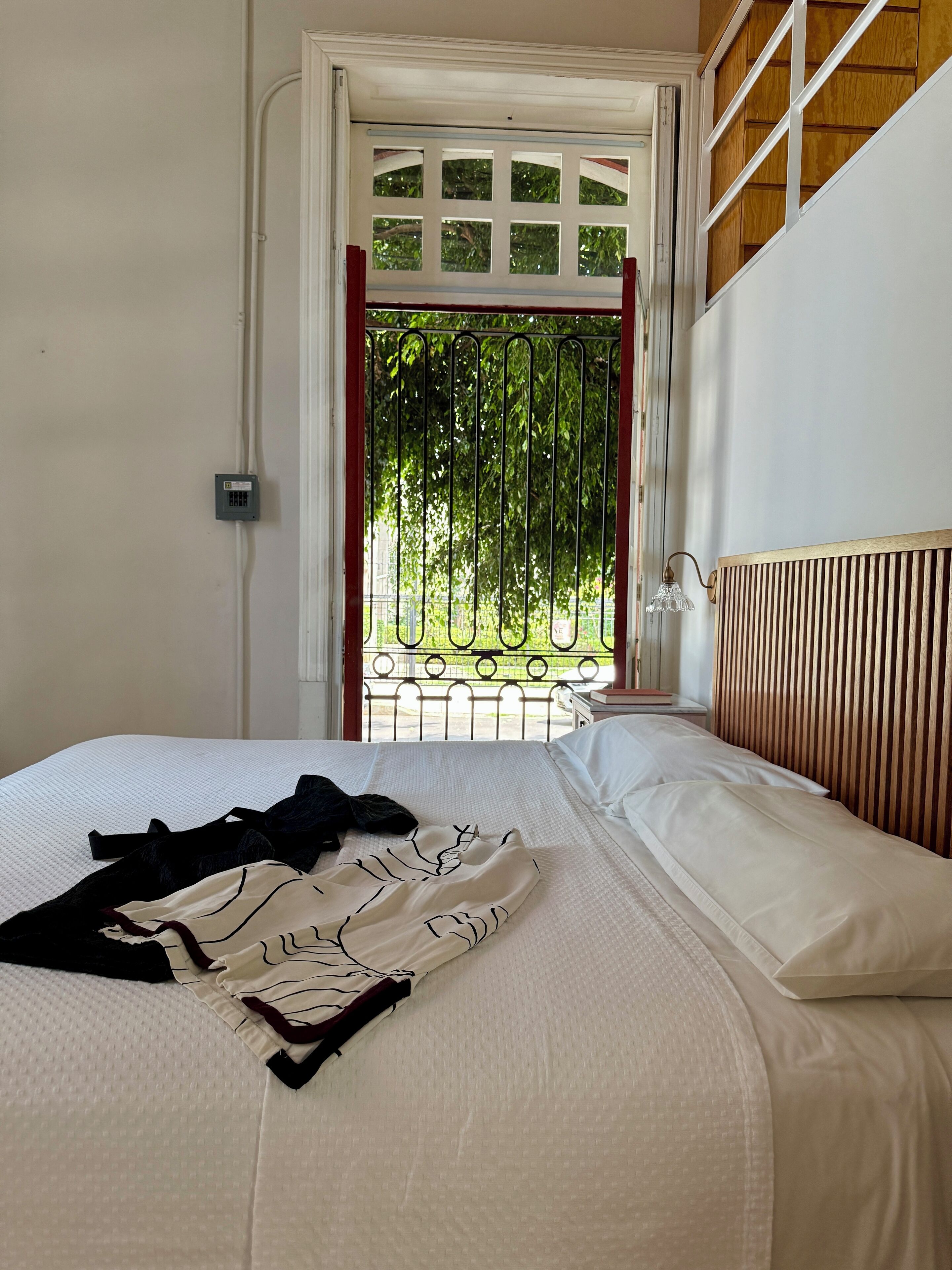 A bedroom with a bed, a radiator, a window with a view of greenery, and a door with a metal gate.