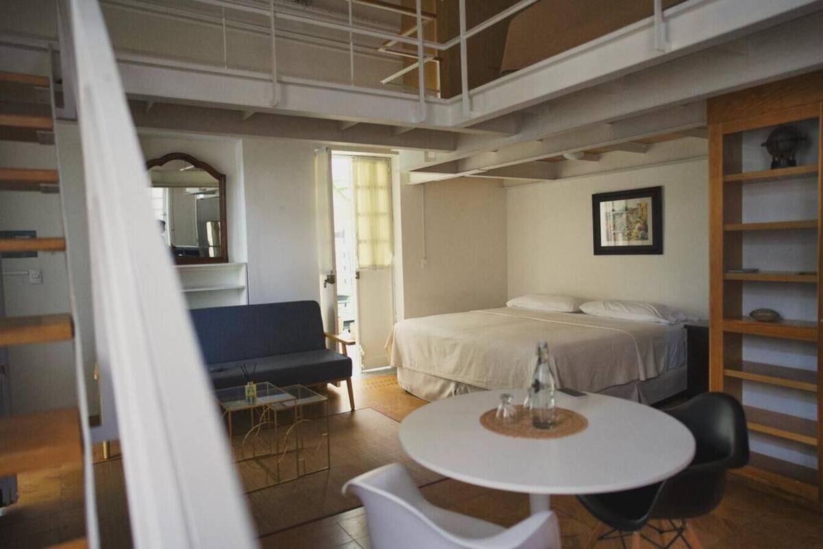 A modern hotel room with a bed, a small table with chairs, a sofa, and a bookshelf.