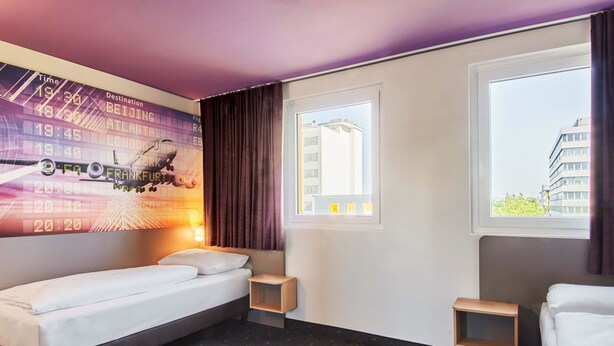 A hotel room with a bed, a small bedside table, a window with a view of buildings, and a wall with a flight information display.
