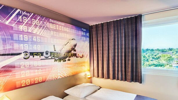 A hotel room with a bed, a window with curtains, and a wall displaying flight information. - B&B Hotel Frankfurt-Airport (Frankfurt)