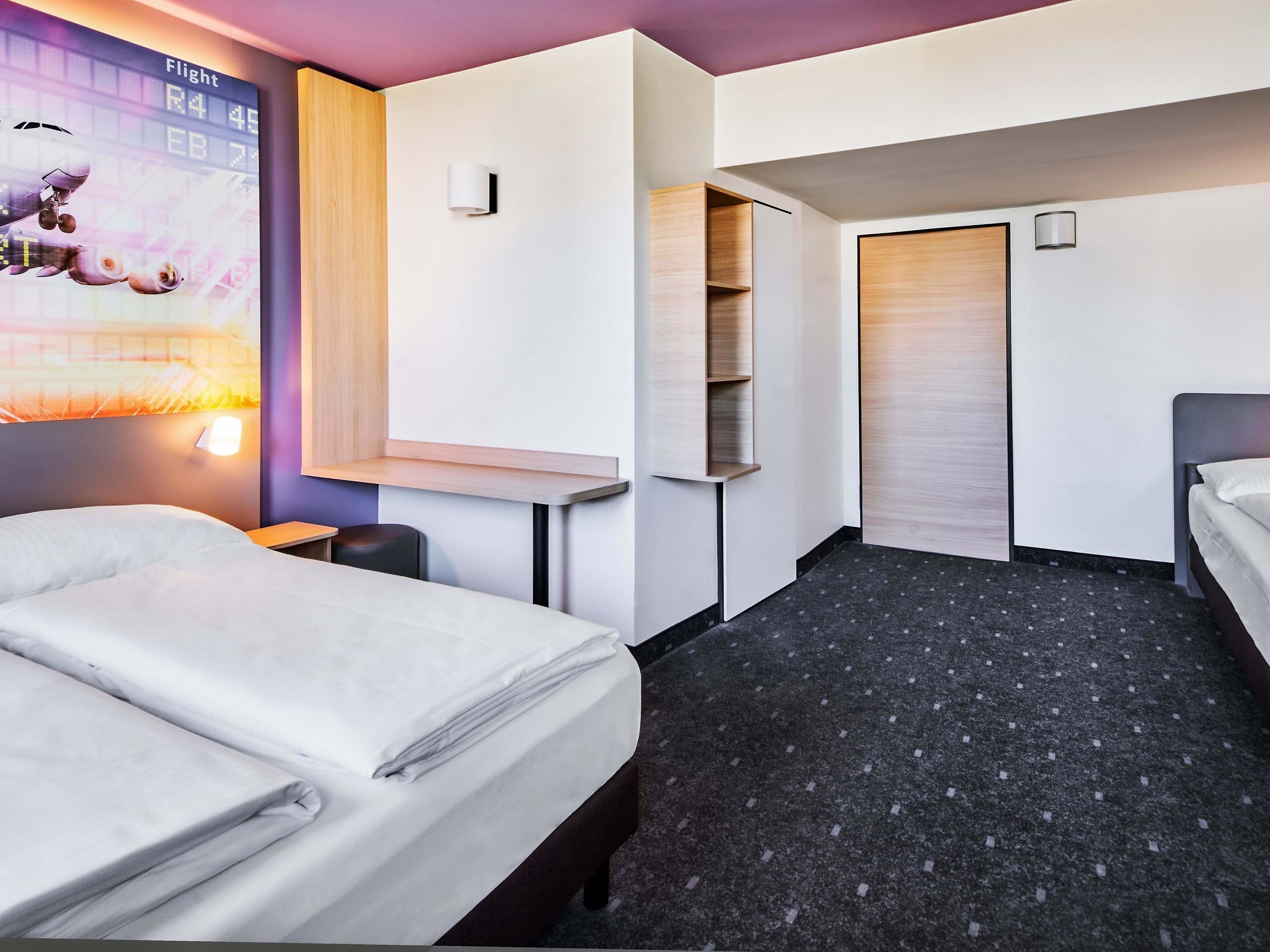 Une chambre d’hôtel avec deux lits, une petite table, une chaise, une armoire et une étagère fixée au mur.