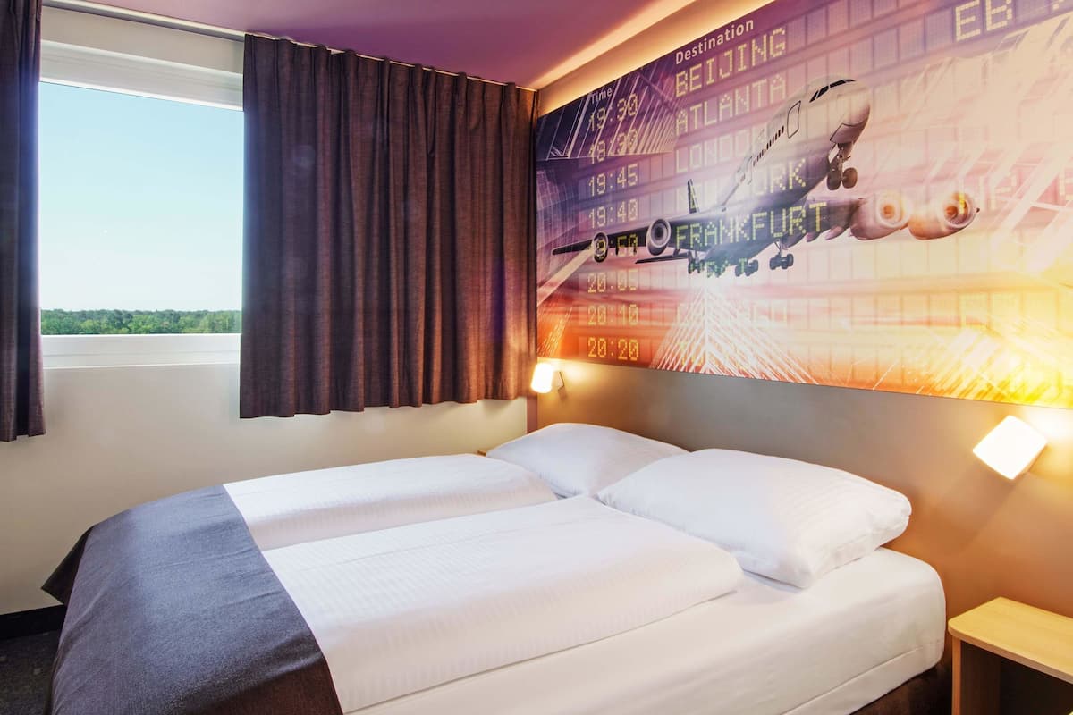 A hotel room with a bed, curtains, a wall clock, and a digital flight information display.