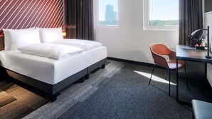 A modern hotel room with a large bed, a desk, and a chair. There are two windows providing views of the city. - B&B Hotel Frankfurt-Airport (Frankfurt)